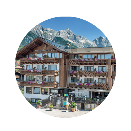 Uriges Hotel mit Holzfassade, vielen Fenstern, und Balkone mit bunten Blumen bestückt, im Hintergrund eine schroffe Gebirgskette und klarem Himmel.