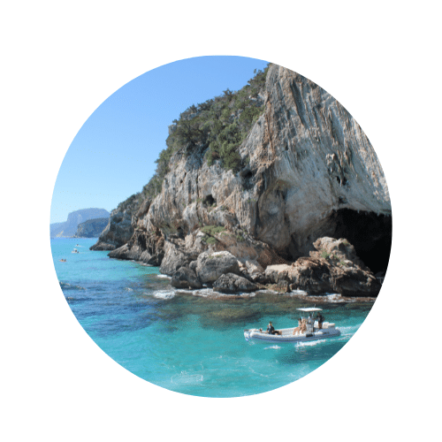 Schroffe Felsvorhänge grün bewachsen mit dunklen Höhleneingängen, darunterliegend türkisblaues Wasser mit einem Schnellboot und Touristen.