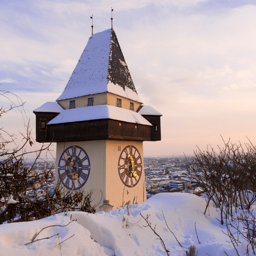 Ein beschneiter Kirchturm in Steiermark. Im Hintergrund der Ort mit verschiedenen Häusern.