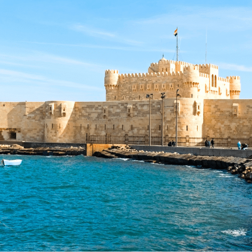 Zu sehen ist die Burg am Hafen von Alexandria mit der ägyptischen Fahne, blauer Himmel und blaues Meer mit einem Fischerboot