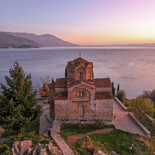 Auf dem Bild zu sehen ist die Kirche des Heiligen Johannes von Kaneo in Ohrid in Nordmazedonien. Links ist ein Tanne und dahinter das Meer mit Bergen.