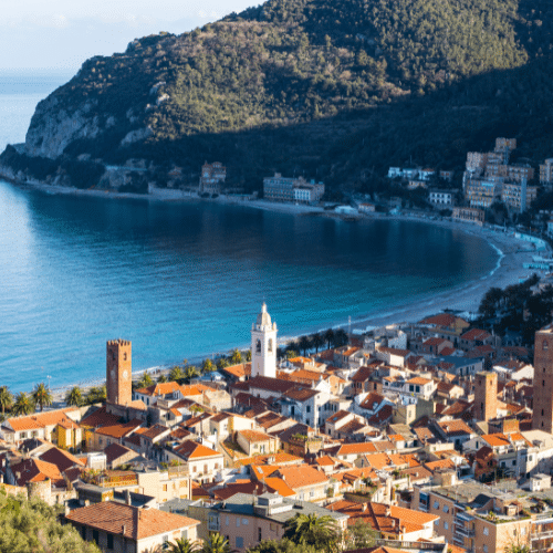 Hier zu sehen ist eine an der Küste liegende Kleinstadt in Savona. Sie beeindruckt über ihre schöne Altstadt recht unten und das blaue Meer. Im Hintergrund ist eine Klippe zu sehen mit grünen Bäumen.