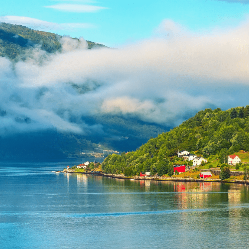 Auf dem Bild zu sehen ist die norwegische Fjordstadt. Links im Bild die Fjorde und rechts die kleine, in der Sonne liegende, Hafenstadt mit roten Häusern. Es ist leicht bewölkt.