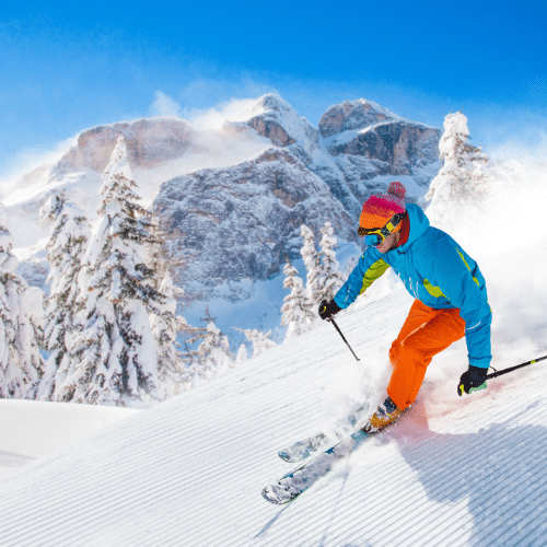 Auf dem Bild zu sehen ist ein Skifahren der die Piste hinab fährt. Im Hintergrund sind in Schnee getauchte Tannen und hohe Berge zu sehen.