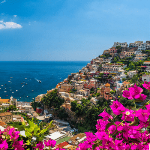 Auf dem Bild zu sehen ist die Amalfiküste in Sorrent. Im Vordergrund sind lilafarbene Blumen, im Hintergrund der Berg mit den bunten Häusern und daneben das blaue Meer.